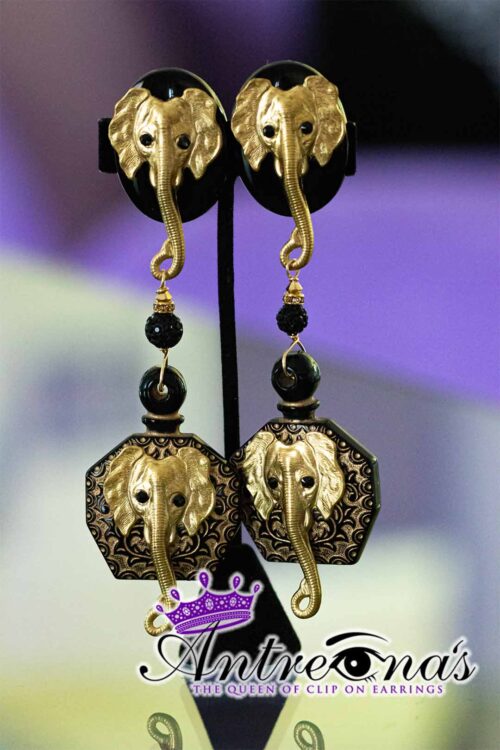 Trunked Majesty is a bold pair of clip-on earrings featuring detailed golden elephant heads with graceful trunks and decorative beads. Hexagonal black and gold patterns frame raised elephant motifs, blending luxury and strength in a regal, elegant design fit for a queen.